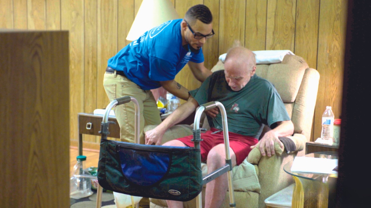 Male caregiver assisting senior with senior health challenges to sit up on a couch.