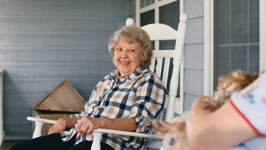 Senior woman client sitting at the balcony looking at her home health aide holding a dog.