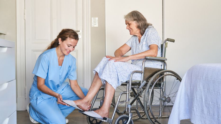 Caregiver helps senior woman in a wheelchair putting a Comfortable Footwear.