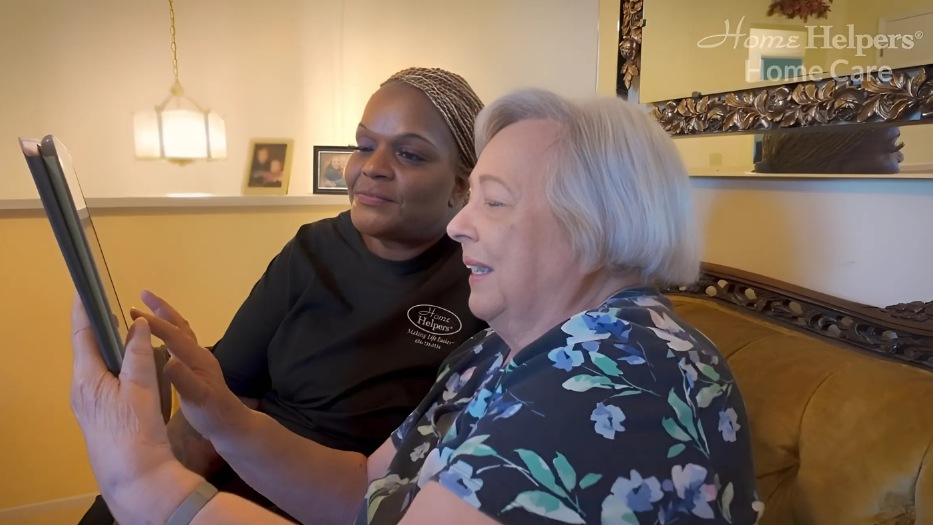 Caregiver sitting beside her senior female senior client who is holding a tablet.