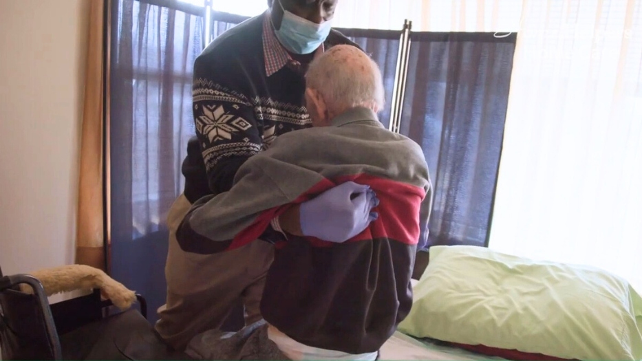 The senior male is being helped by Home Helpers caregiver from transferring from wheelchair to bed.