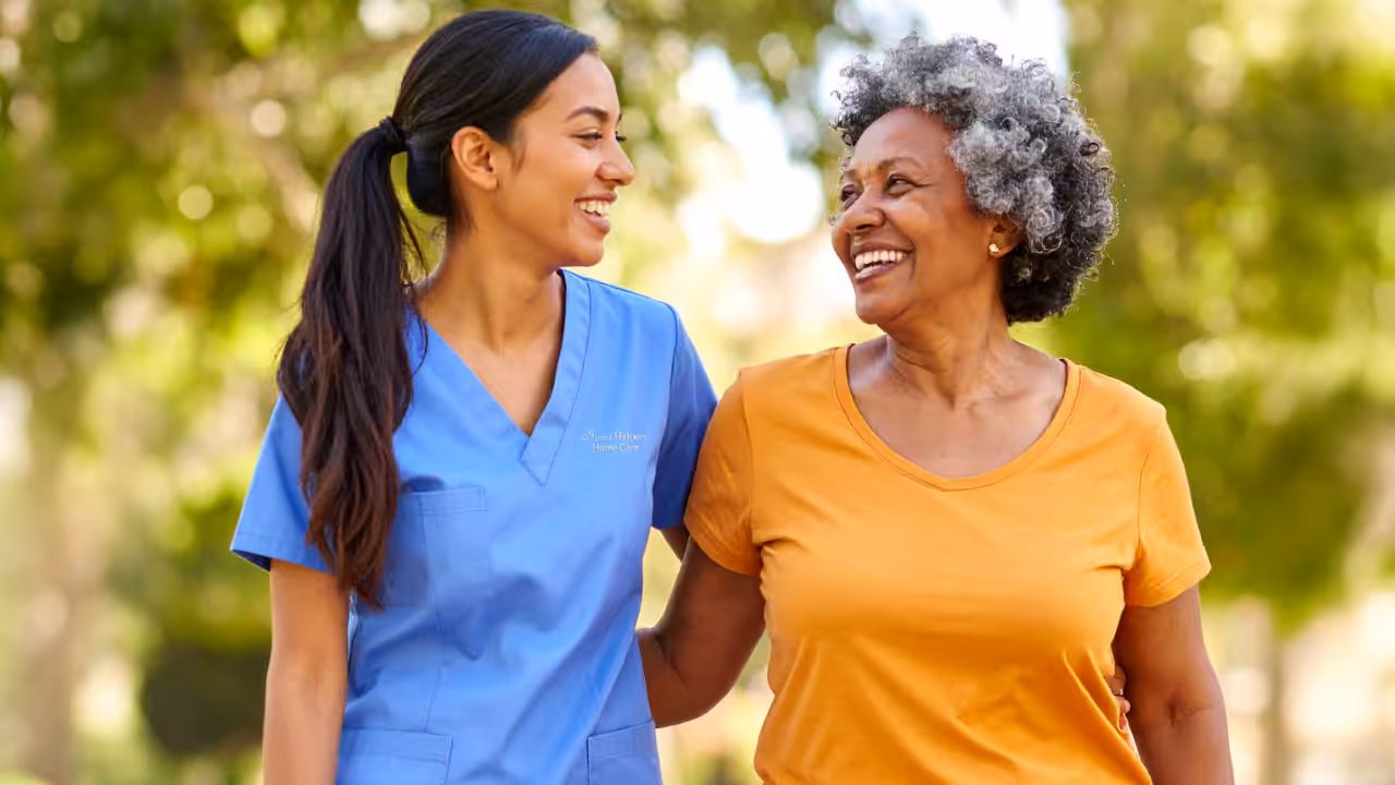 A young woman in blue scrubs and an older woman in an orange shirt smile at each other while walking outside, arm in arm, with green trees blurred in the background.