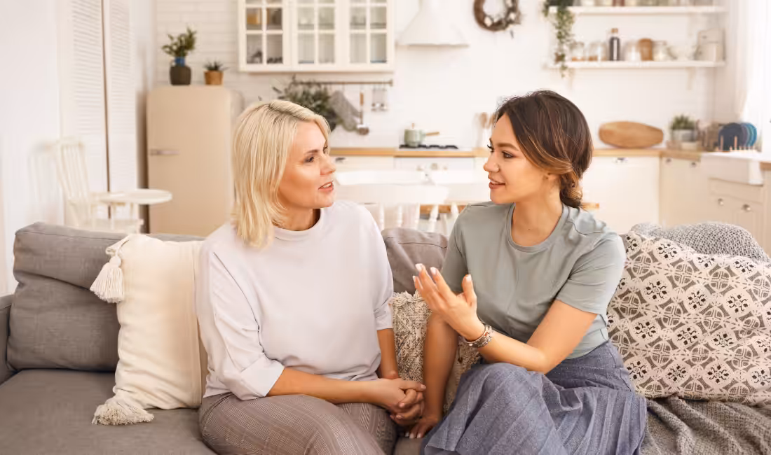 Two women sit on a gray couch in a cozy, bright kitchen, having a friendly conversation. One woman gestures with her hand while the other listens attentively. Pillows and home decor create a welcoming atmosphere.