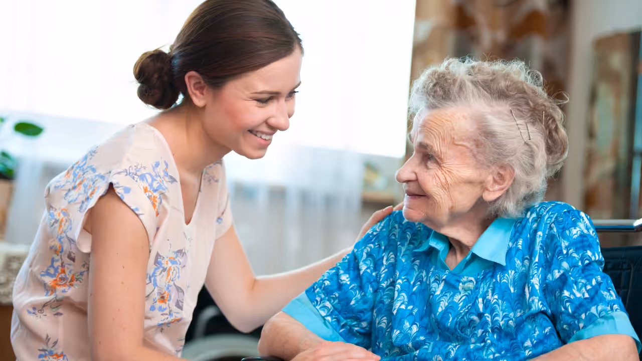 A young woman smiles warmly while gently touching the shoulder of an elderly woman in a wheelchair. They are indoors and appear to be enjoying a friendly conversation.