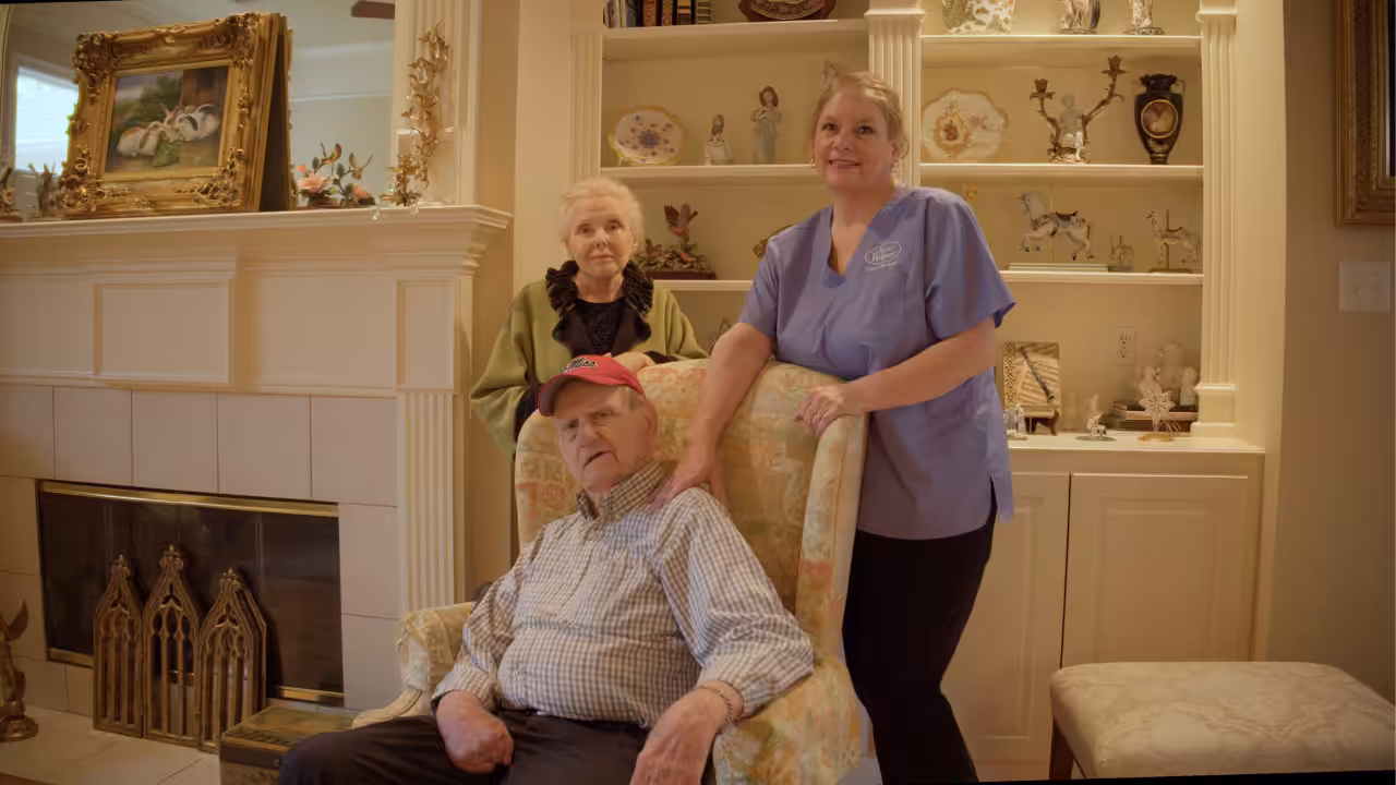 An elderly man sits in an armchair, a caregiver in blue scrubs stands beside him, and an older woman stands in the background. They are in a warmly decorated living room with shelves and a fireplace.