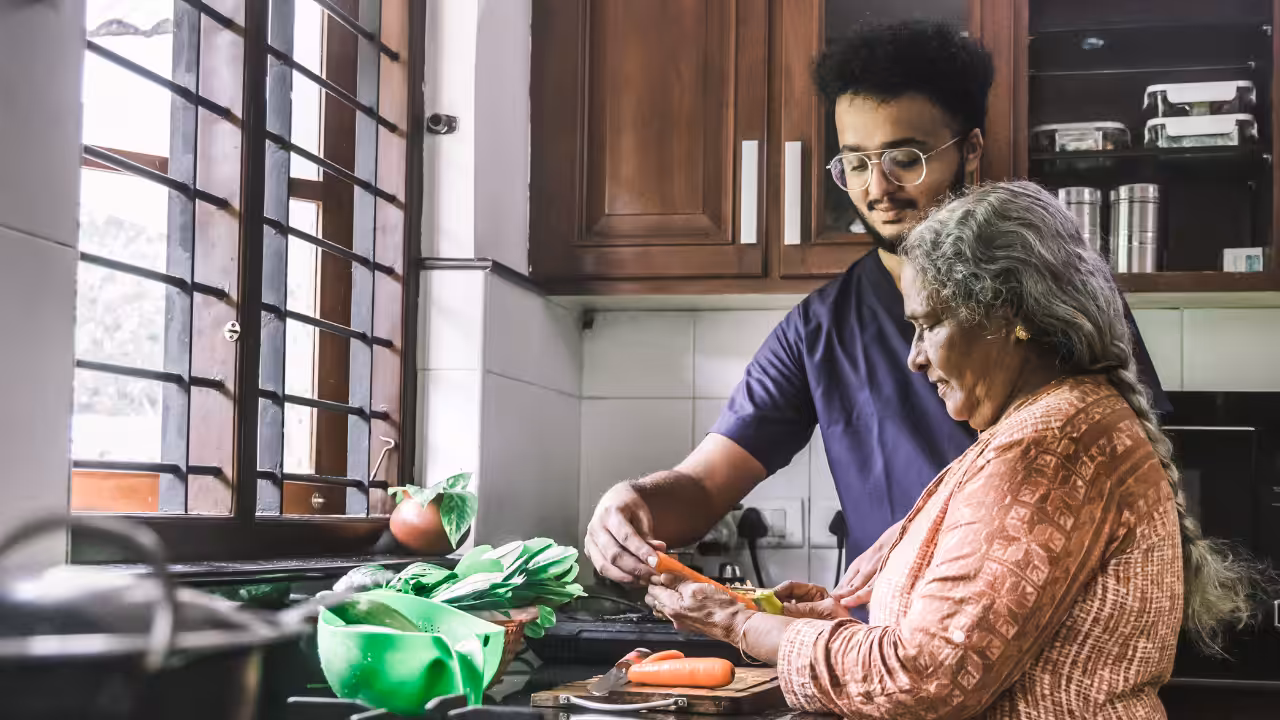 A young man helps an elderly woman peel vegetables in a bright kitchen. They stand together by the counter near a window with sunlight streaming in. Fresh produce and utensils are visible on the counter.