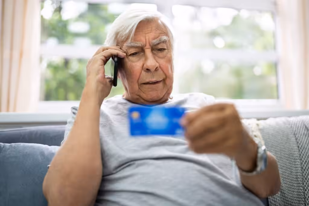 An elderly man sits on a couch, holding a phone to his ear and looking thoughtfully at a credit card, appearing concerned—an image that highlights the growing issue of elder fraud. Bright window light fills the background.