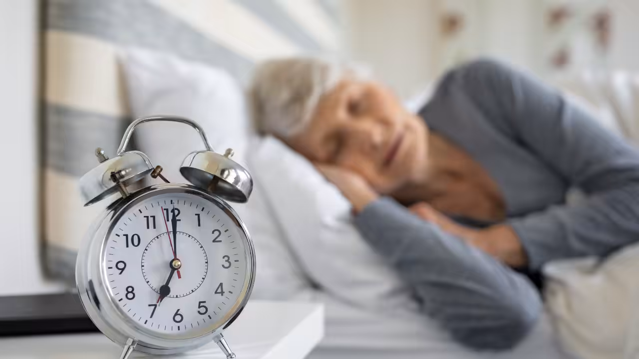 An elderly person sleeps peacefully in bed, with a silver analog alarm clock showing 8:20 on a nearby white nightstand in the foreground.