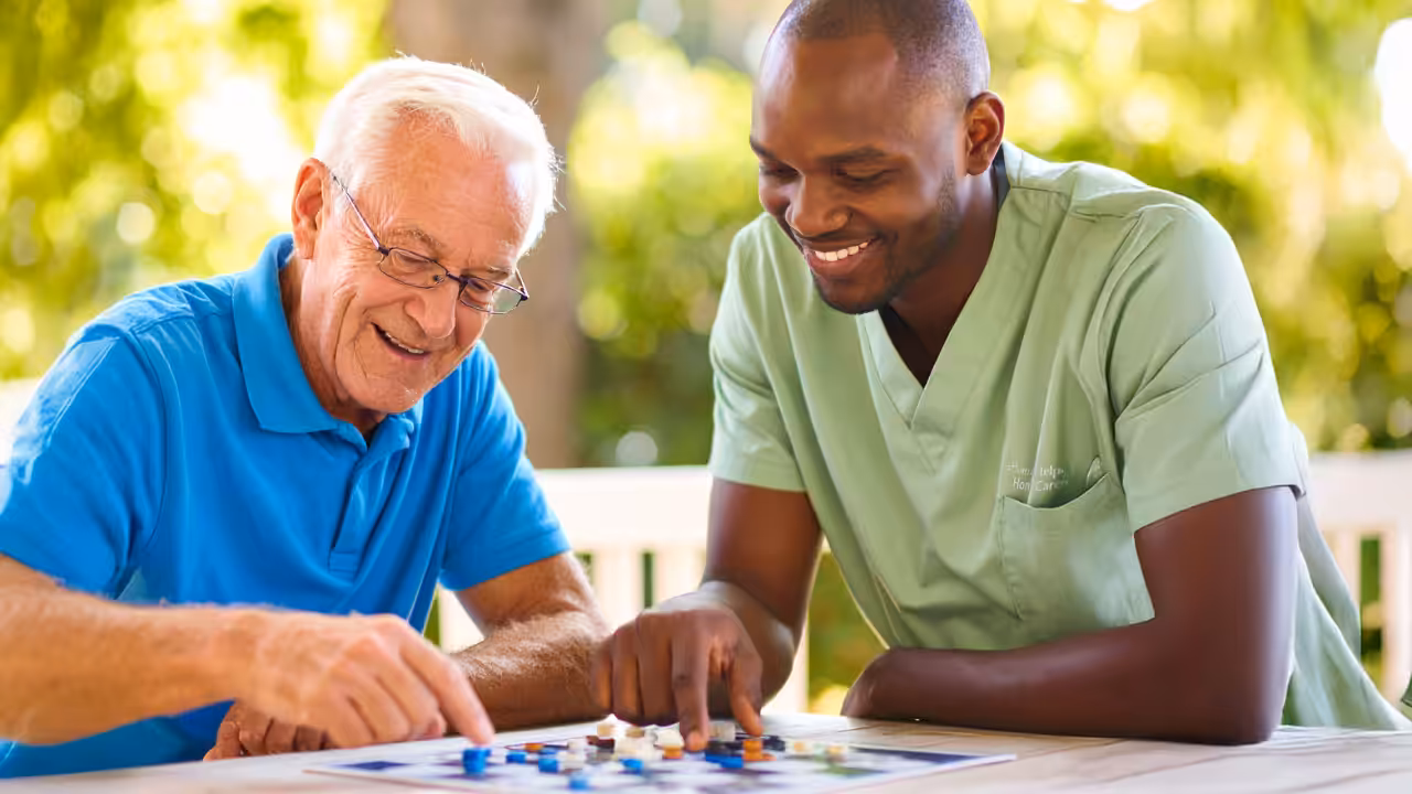 An older man and a younger man sit at a table outdoors, smiling and playing a board game together. The younger man is wearing light green scrubs, suggesting he is a caregiver or healthcare worker.