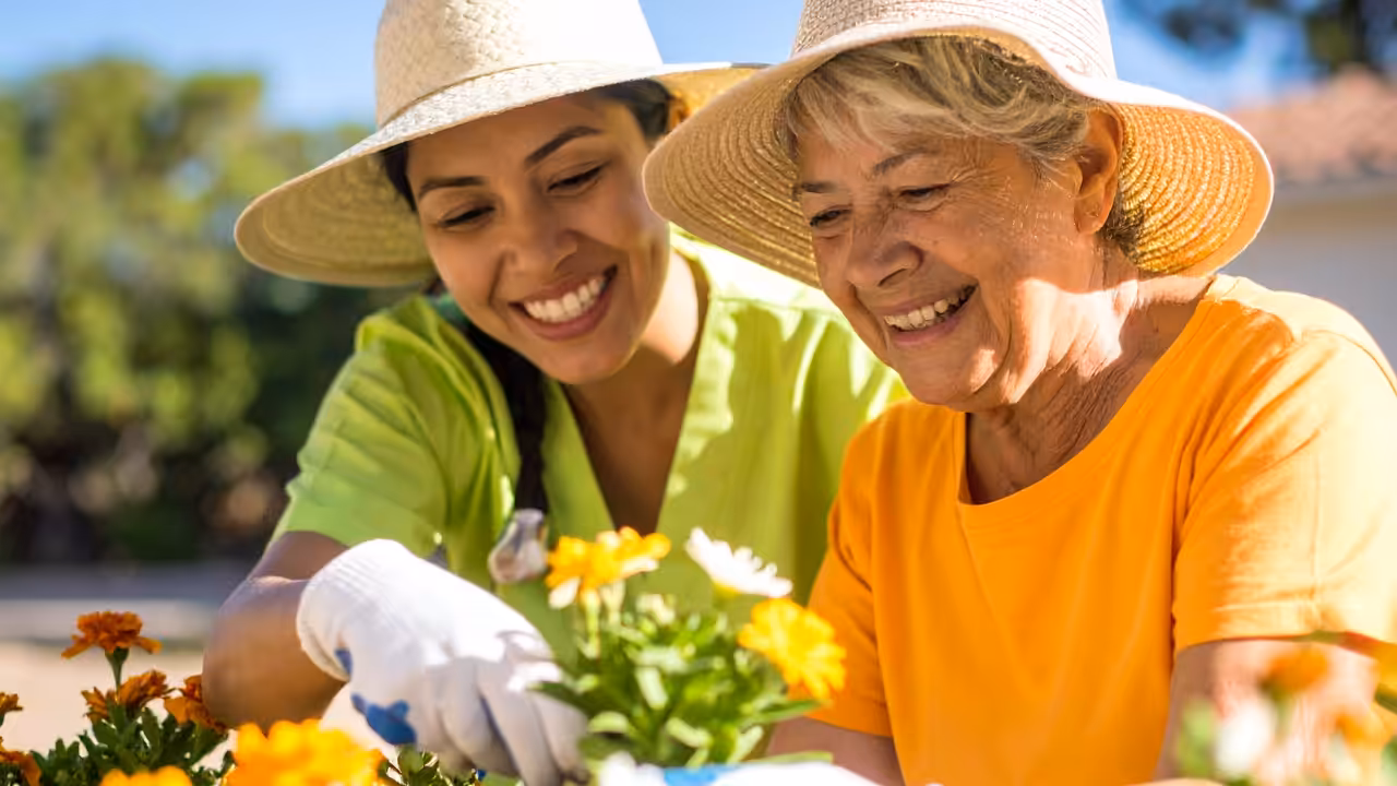Two women, one younger and one older, both wearing sun hats and gloves, smile as they garden together, tending to yellow and white flowers outdoors on a sunny day.
