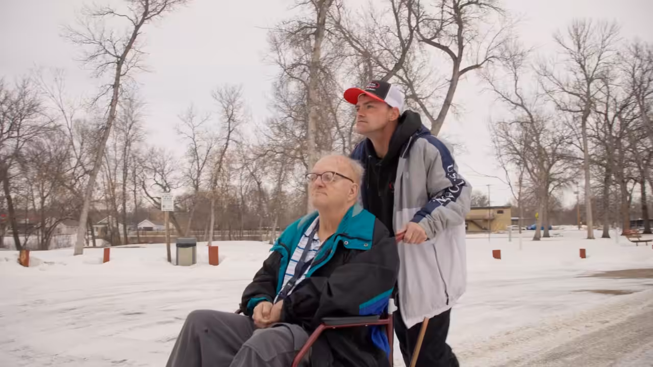 A man in a wheelchair is being pushed by another man outdoors on a snowy day. Both are dressed warmly, and bare trees are visible in the background. The scene appears calm and peaceful.