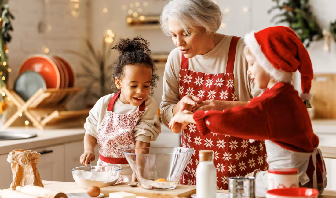 An older woman and two young children, all in aprons, bake together in a festive kitchen. One child wears a Santa hat, and they are cracking eggs into a bowl, smiling and enjoying the holiday activity.