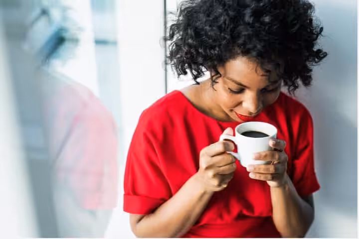 A woman with curly hair wearing a red top sits by a window, smiling and holding a white cup of coffee with both hands.