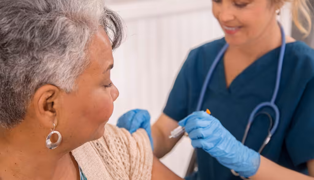 A nurse wearing blue scrubs and gloves prepares to give a vaccine injection to an older woman with short gray hair, who is sitting with her sleeve rolled up. Both appear calm in a clinical setting.