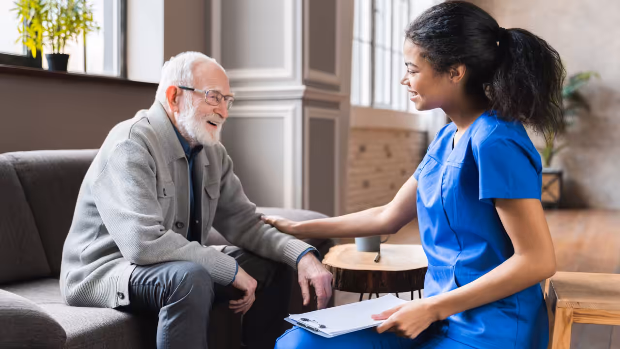 A nurse in blue scrubs smiles and talks with an elderly man sitting on a couch, gently touching his arm and holding a clipboard, in a bright, comfortable room.