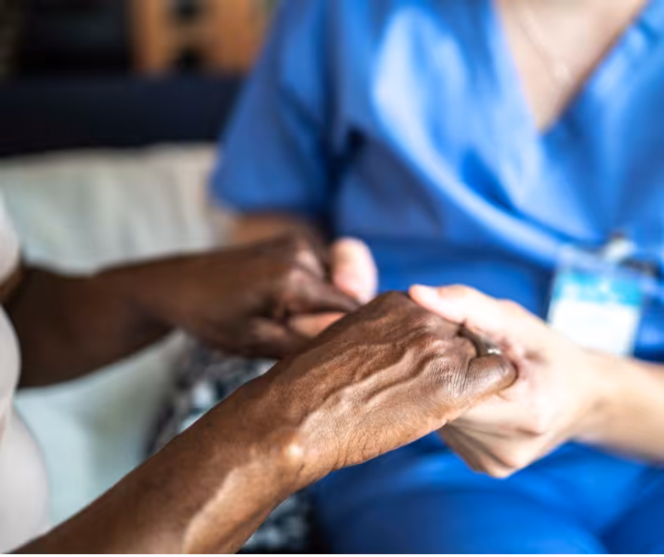 A healthcare worker in scrubs gently holds the hands of an elderly person, offering comfort and support. The image focuses on their hands, symbolizing care and compassion.