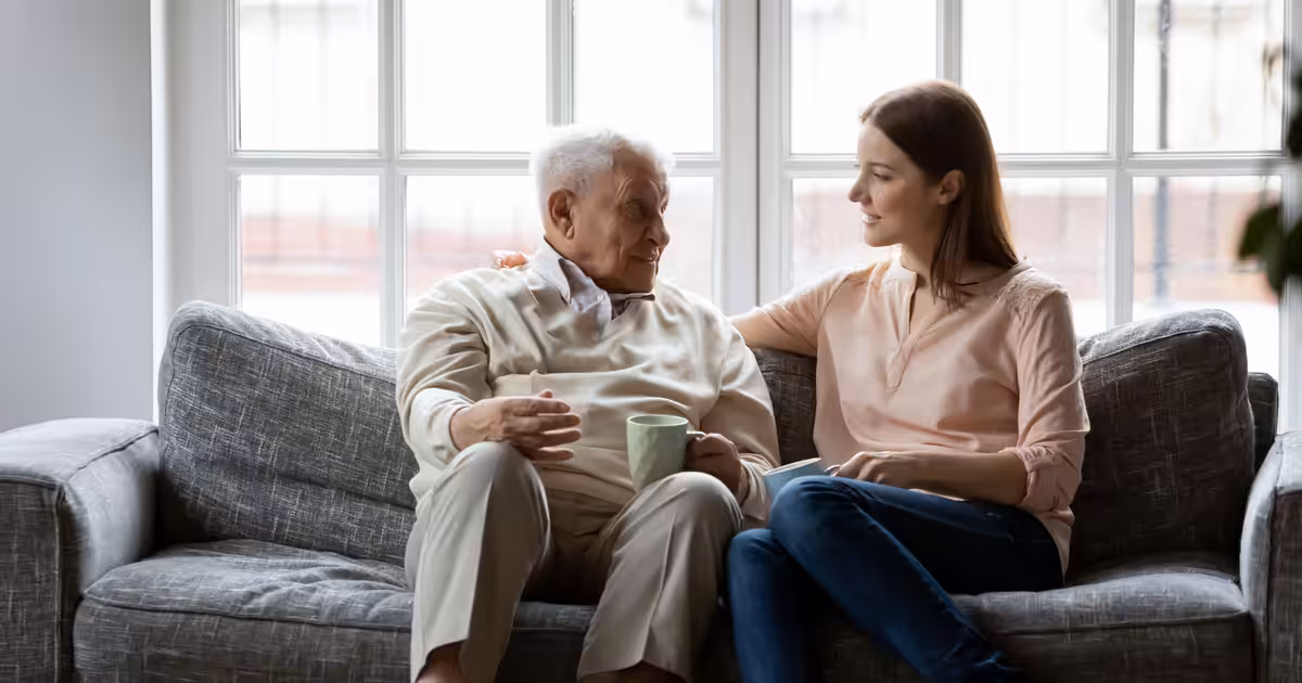 A young woman and an elderly man sit together on a gray sofa in a bright room. The woman has her arm around the man, who is holding a mug, and they appear to be having a warm, engaged conversation.