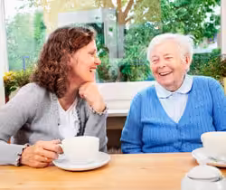A younger woman and an older woman sit at a wooden table, smiling and laughing together while enjoying hot drinks. A window with green trees is visible in the background.