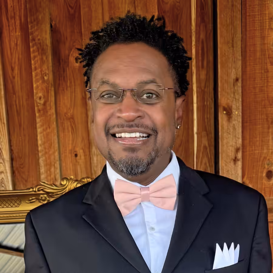 A man in a black suit, white shirt, and pink bow tie smiles at the camera. He is wearing glasses and standing in front of a wooden background with decorative golden accents.