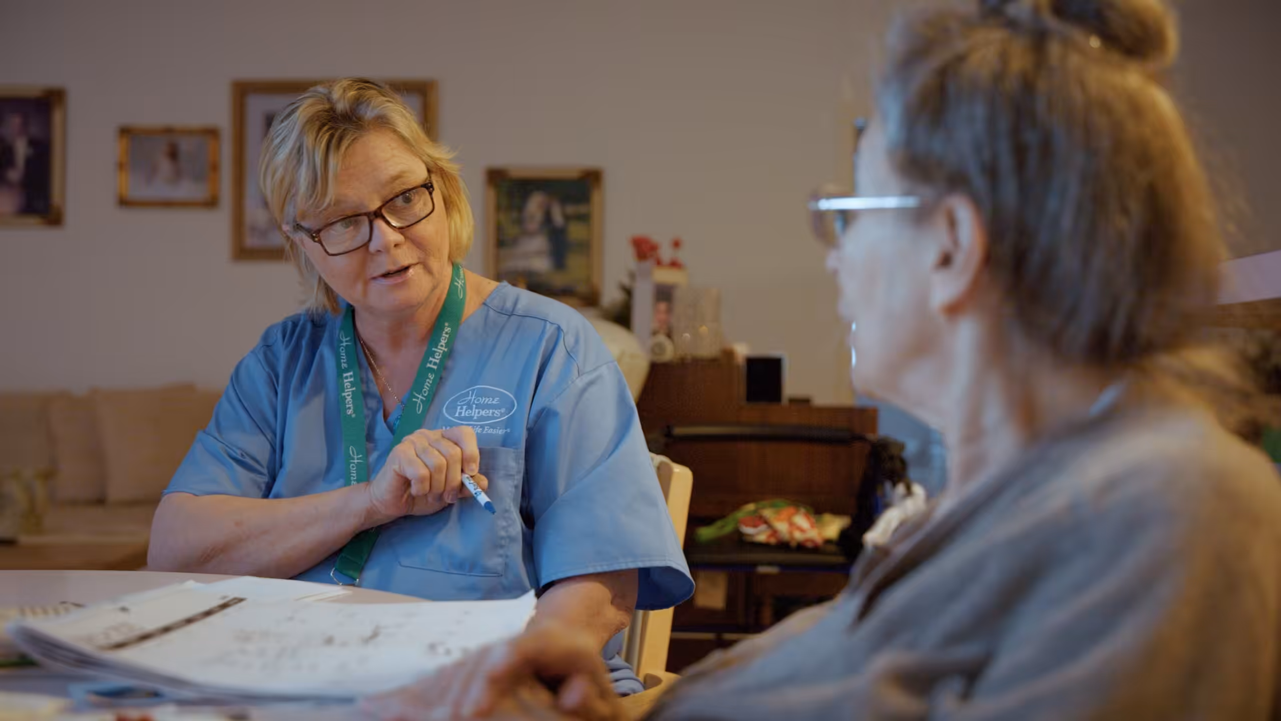 A nurse in blue scrubs speaks to an elderly woman at a table, holding a pen and reviewing documents, in a warmly lit room with framed photos on the wall in the background.