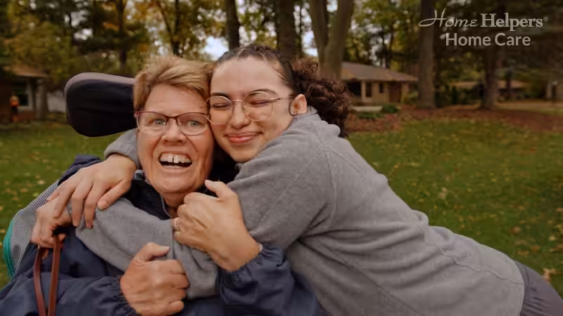 An elderly woman in a wheelchair smiles as a young woman hugs her from the side outside in a park-like setting. Both look happy. The words “Home Helpers Home Care” are written in the top right corner.