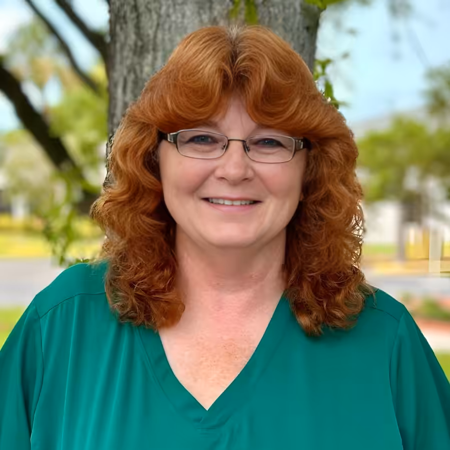 A woman with wavy red hair and glasses smiles while standing outdoors in front of a tree. She is wearing a green top, and the background shows blurred trees and buildings.