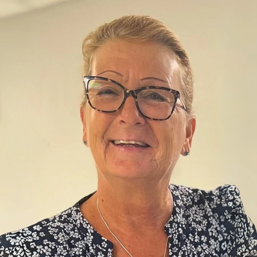 A smiling older woman with short, light hair and glasses, wearing a dark floral top and a necklace, stands in front of a plain light background.