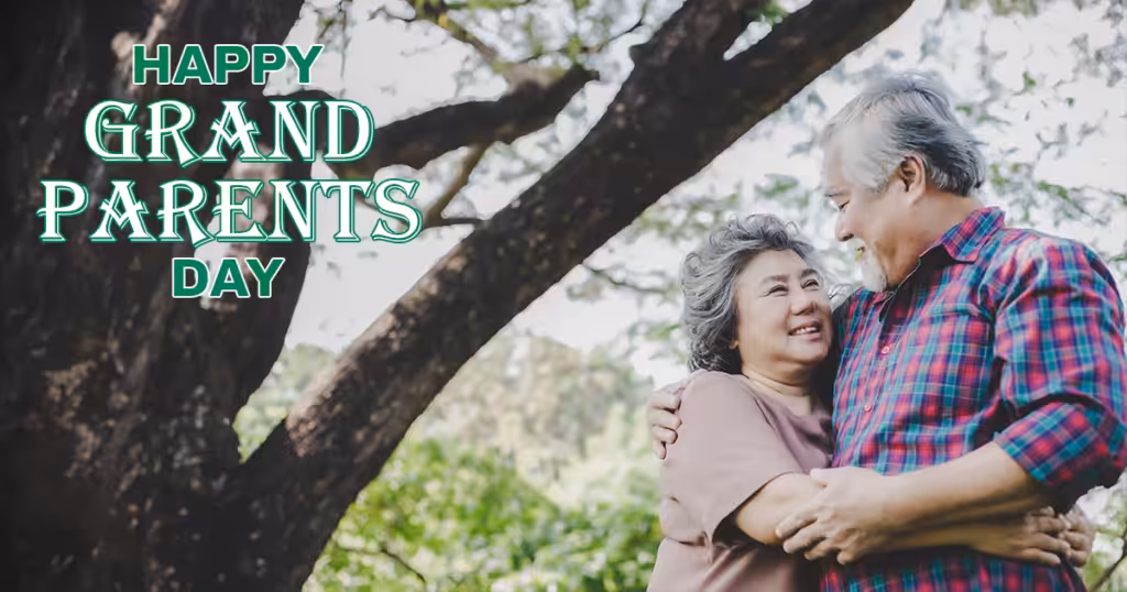 An elderly couple smiles and embraces under a large tree in a park. The text Happy Grandparents Day appears in bold green letters on the left side of the image.