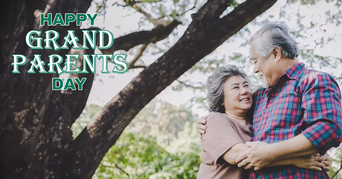 An elderly couple smiles and embraces under a large tree in a park. The text Happy Grandparents Day appears in bold green letters on the left side of the image.
