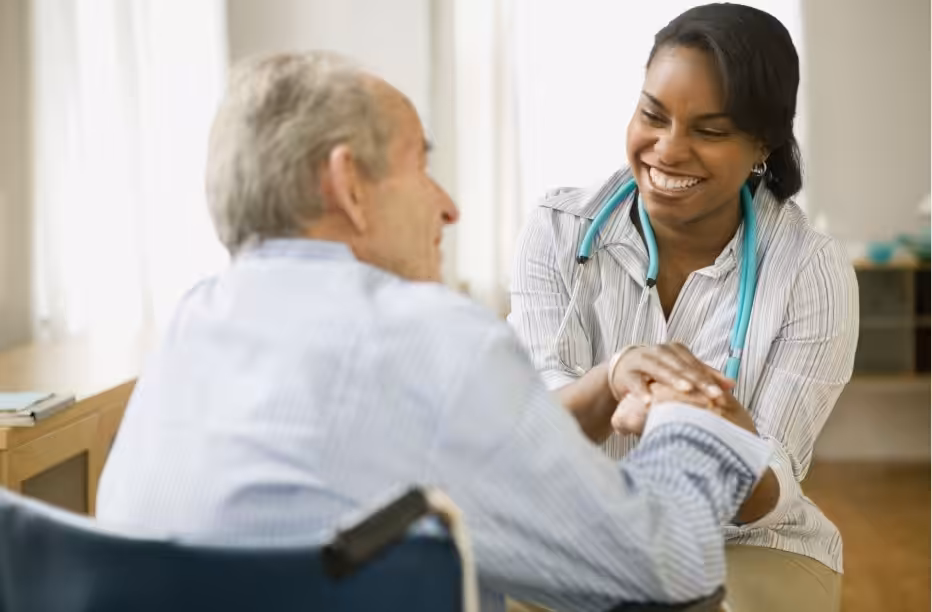 A smiling healthcare professional with a stethoscope holds the hand of an elderly man in a wheelchair, offering comfort and support in a bright room.