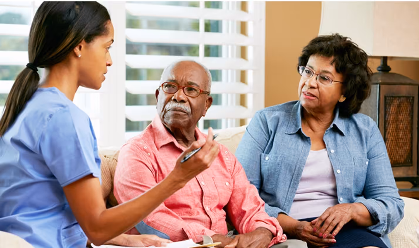 A nurse or caregiver talks with an elderly man and woman sitting on a couch, discussing paperwork in a bright, comfortable room. The man looks attentive while the woman listens closely.