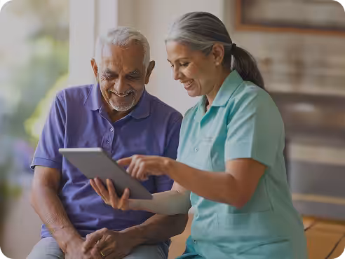 A smiling elderly man sits next to a caregiver in a teal uniform, both looking at and interacting with a tablet device in a bright room.