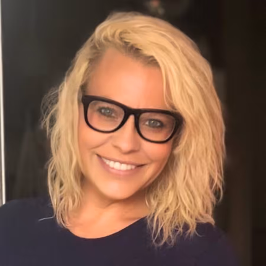 A woman with wavy blonde hair and black glasses smiles at the camera. She is wearing a dark top and is standing indoors with a blurred background.