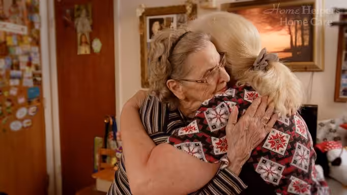 Two elderly women warmly embrace each other in a cozy, decorated room, expressing comfort and affection. Framed photos and a sunset picture are visible in the background.
