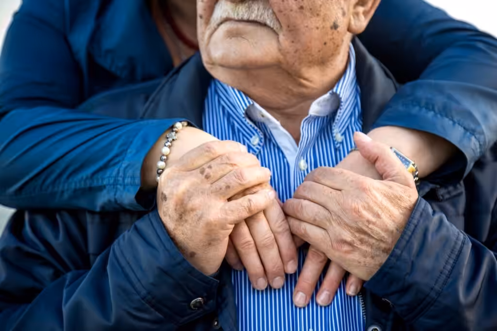 An elderly man wearing a blue striped shirt and jacket is gently embraced from behind by another person, whose hands rest on his chest, showing comfort and support. Both hands feature age spots and jewelry.