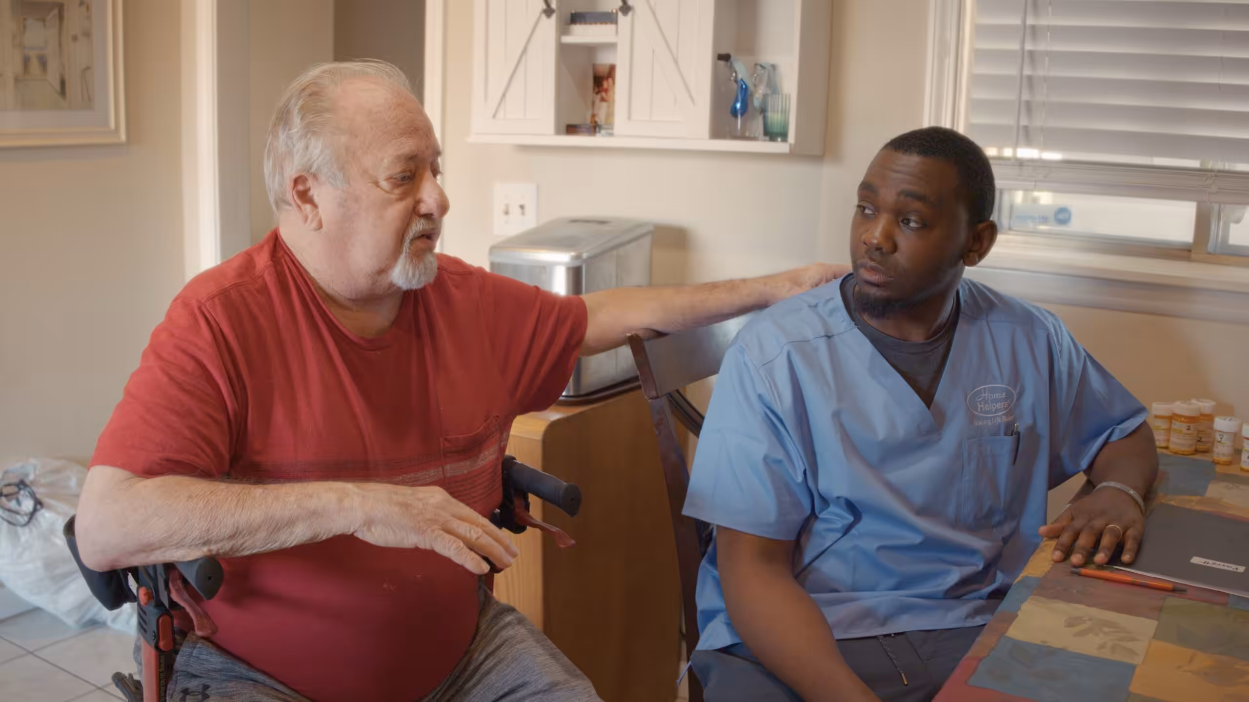 An elderly man in a red shirt sits and talks with a younger man in blue scrubs at a table in a home setting. The elderly man has his hand on the other’s shoulder, and they both appear engaged in conversation.