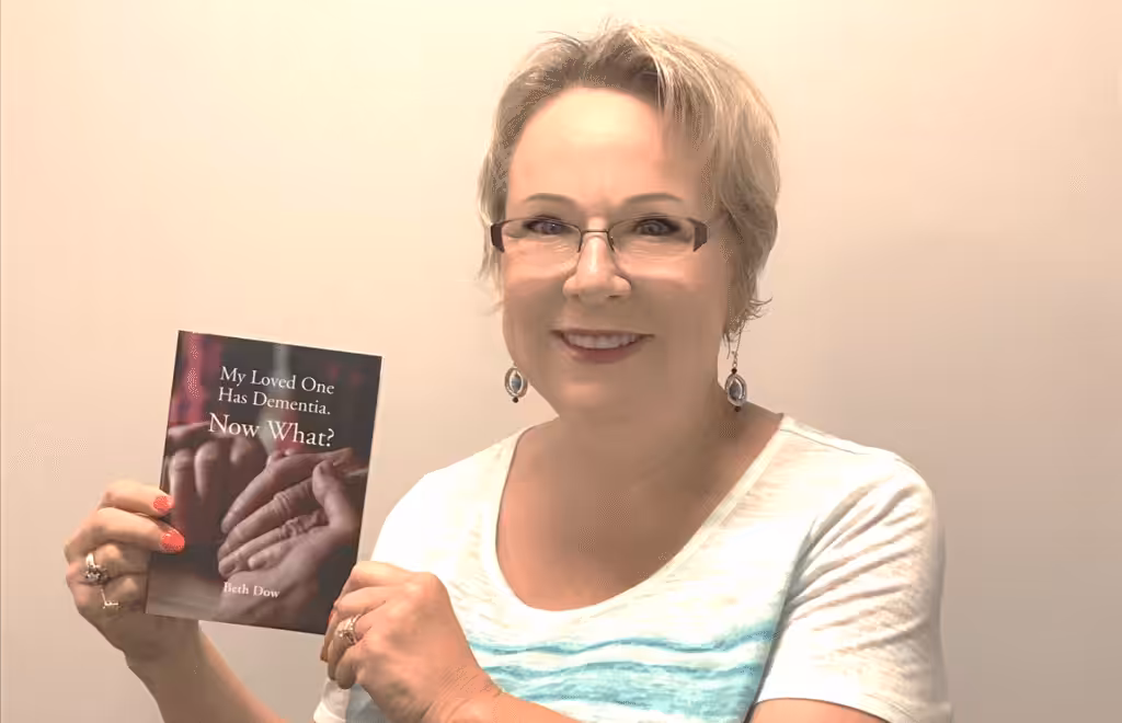 A smiling woman with short blonde hair and glasses holds up a book titled My Loved One Has Dementia. Now What? The cover shows two hands touching gently. The woman is wearing a light-colored shirt and earrings.