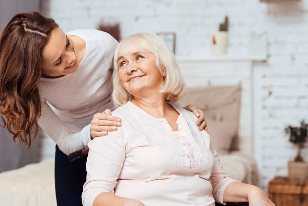 A young woman stands behind an older woman sitting in a chair, gently placing her hands on the older womans shoulders. Both are smiling warmly at each other in a cozy, well-lit room.