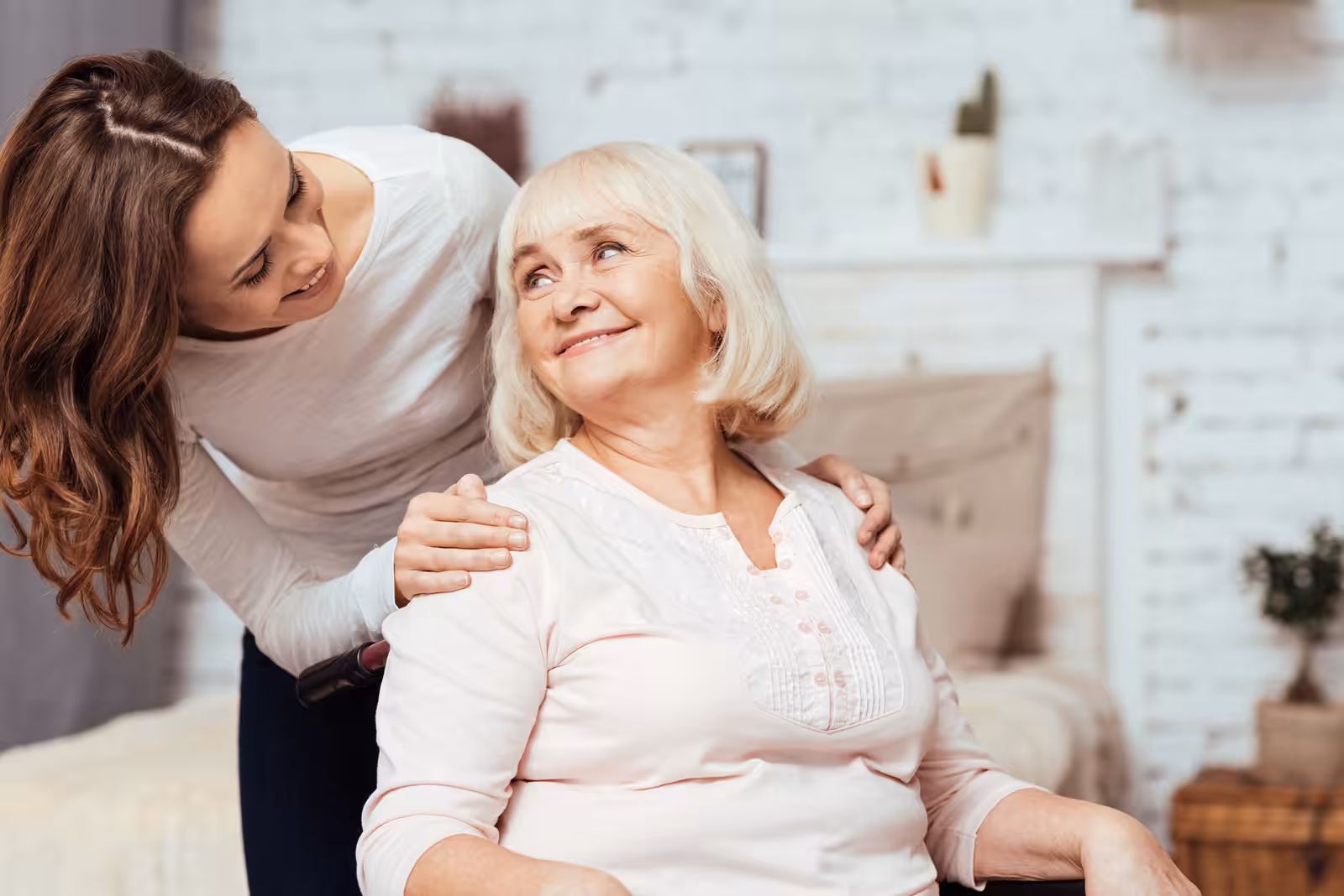 A young woman stands behind an older woman sitting in a chair, gently placing her hands on the older womans shoulders. Both are smiling warmly at each other in a cozy, well-lit room.