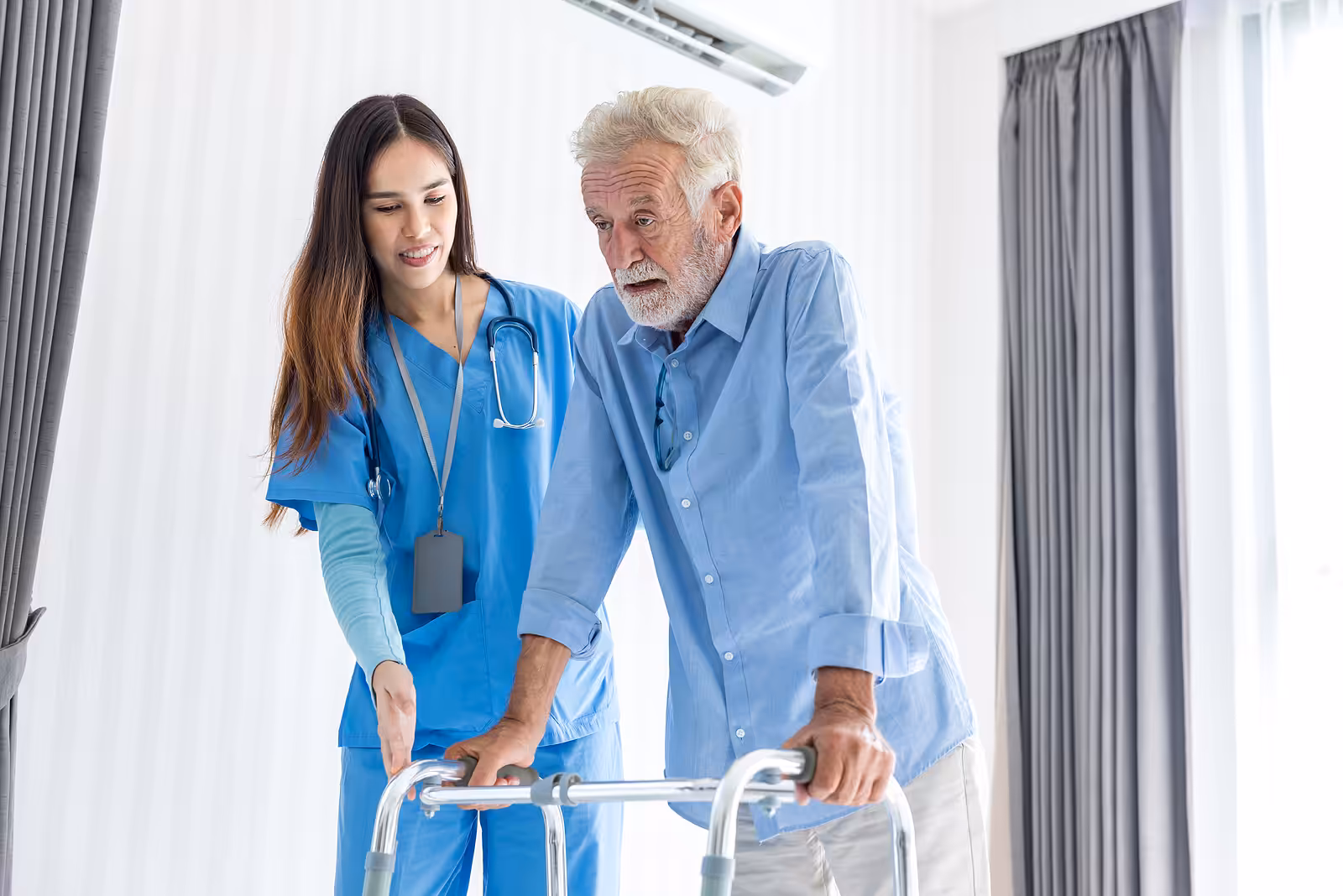 A young female healthcare worker assists an elderly man with gray hair as he uses a walker in a bright room with large windows and curtains.