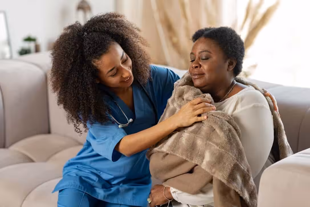 A nurse in blue scrubs comforts a woman wrapped in a blanket who appears to be upset and crying, sitting together on a beige couch in a cozy living room.