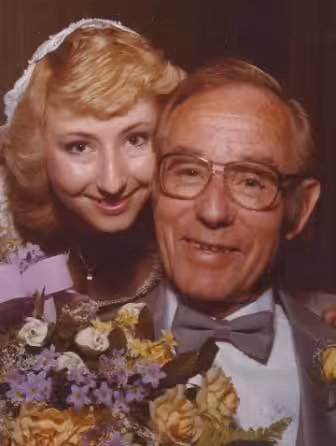 A smiling woman in a wedding dress and veil poses closely with an older man in glasses and a bow tie, both holding a bouquet of colorful flowers with yellow, white, and purple blooms.