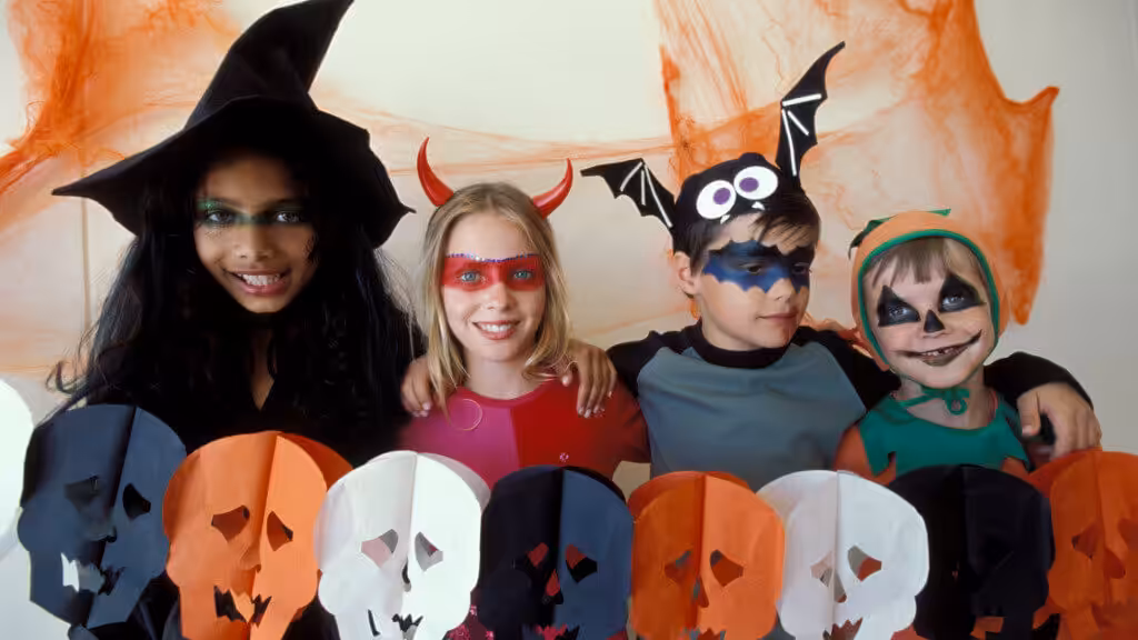 Four children in colorful Halloween costumes (a witch, devil, bat, and sprite) smile in front of orange decorations, holding a garland of black, white, and orange paper skulls.