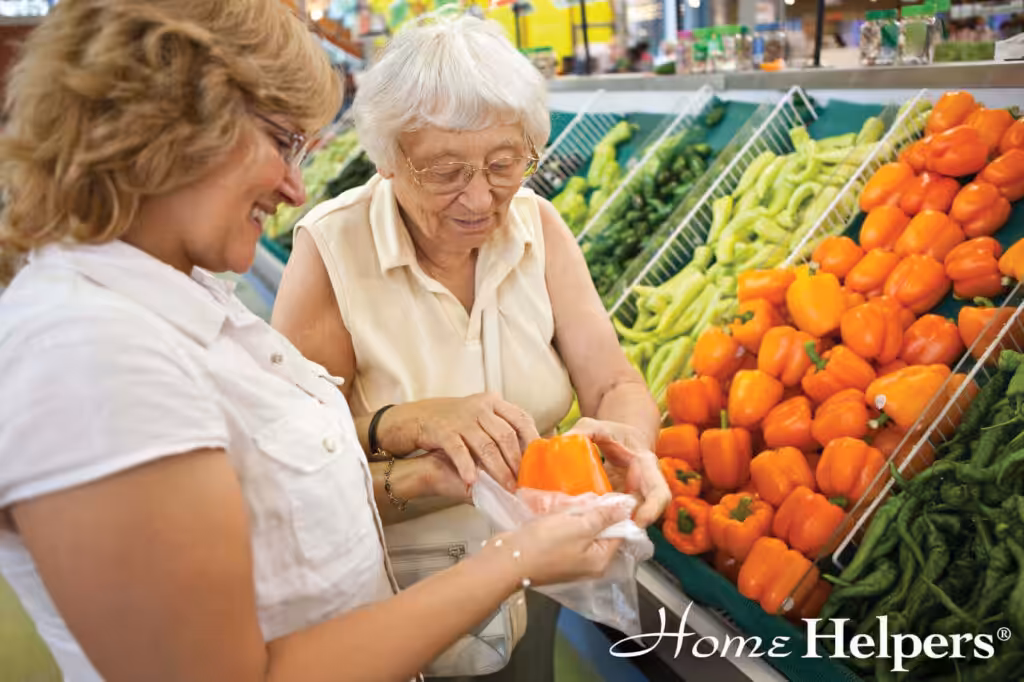 An elderly woman and a younger woman smiling while selecting orange bell peppers at a grocery store produce section. The Home Helpers logo is visible in the corner.