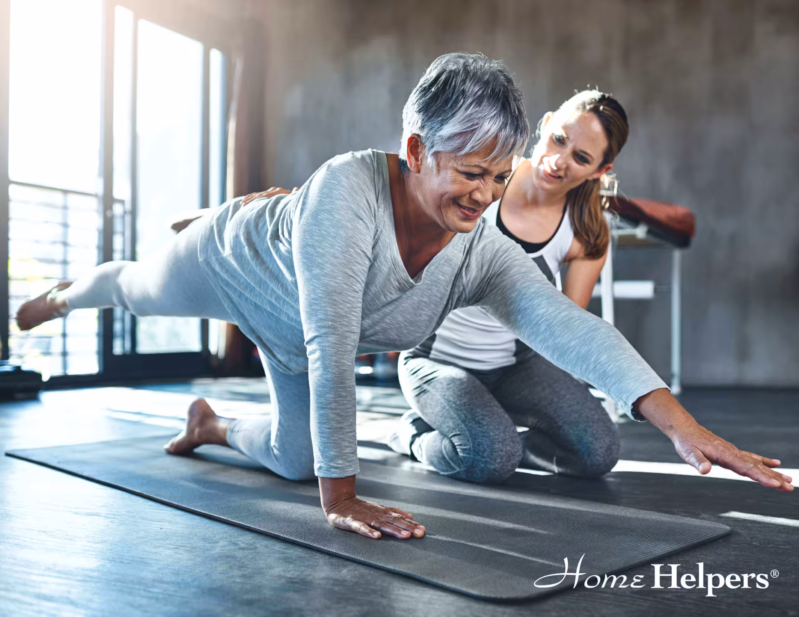 An older woman in workout clothes does a bird-dog exercise on a mat, extending one arm and the opposite leg, while a younger woman kneels beside her and offers encouragement. The Home Helpers logo appears in the corner.
