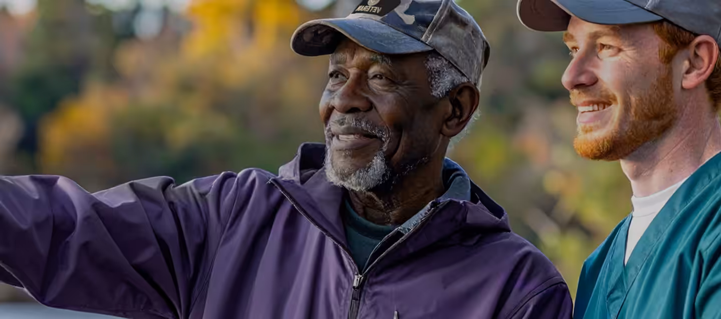 Two men outdoors, one older and one younger, both smiling. The older man wears a purple jacket and a cap, and is pointing at something off-camera. The younger man has a beard and wears a cap and green shirt. Trees blur in the background.