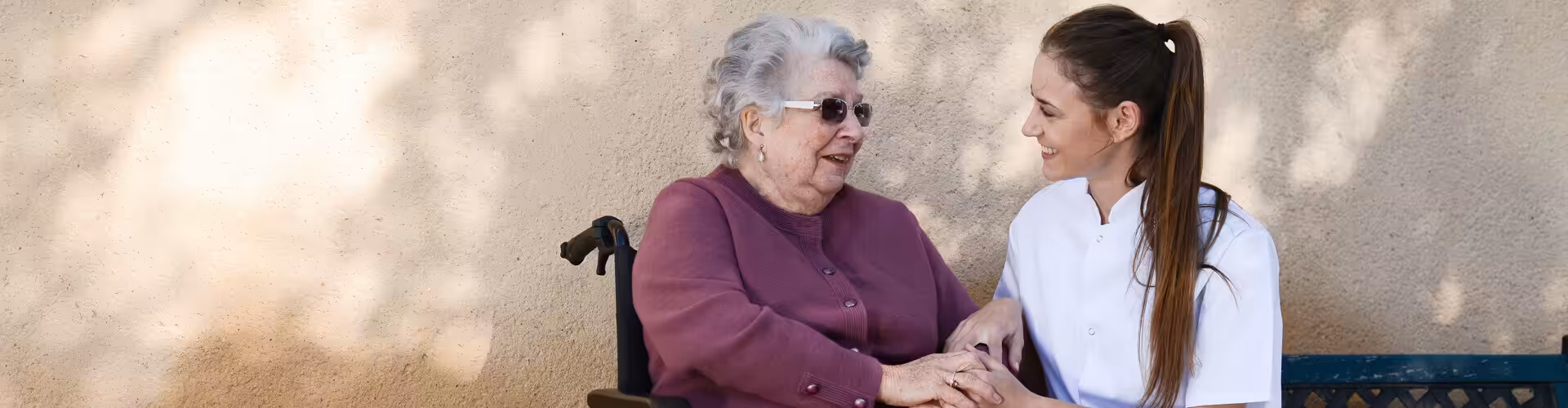 A smiling elderly woman in a wheelchair holds hands with a young female caregiver in a white uniform, both sitting and talking outside against a sunlit beige wall.