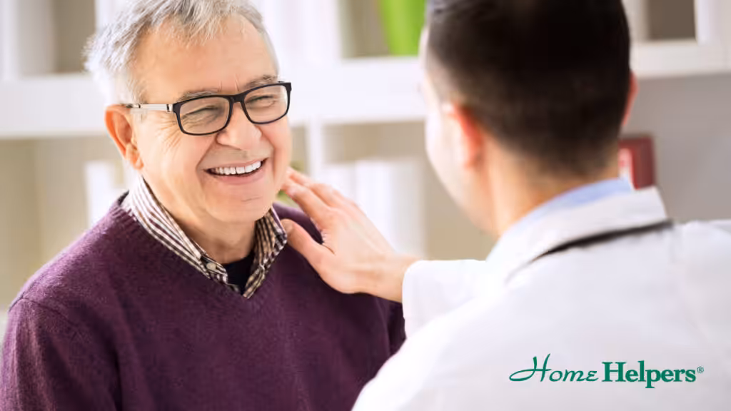 A smiling older man wearing glasses and a purple sweater sits while a healthcare professional in a white coat gently touches his shoulder. The Home Helpers logo appears in the bottom right corner.