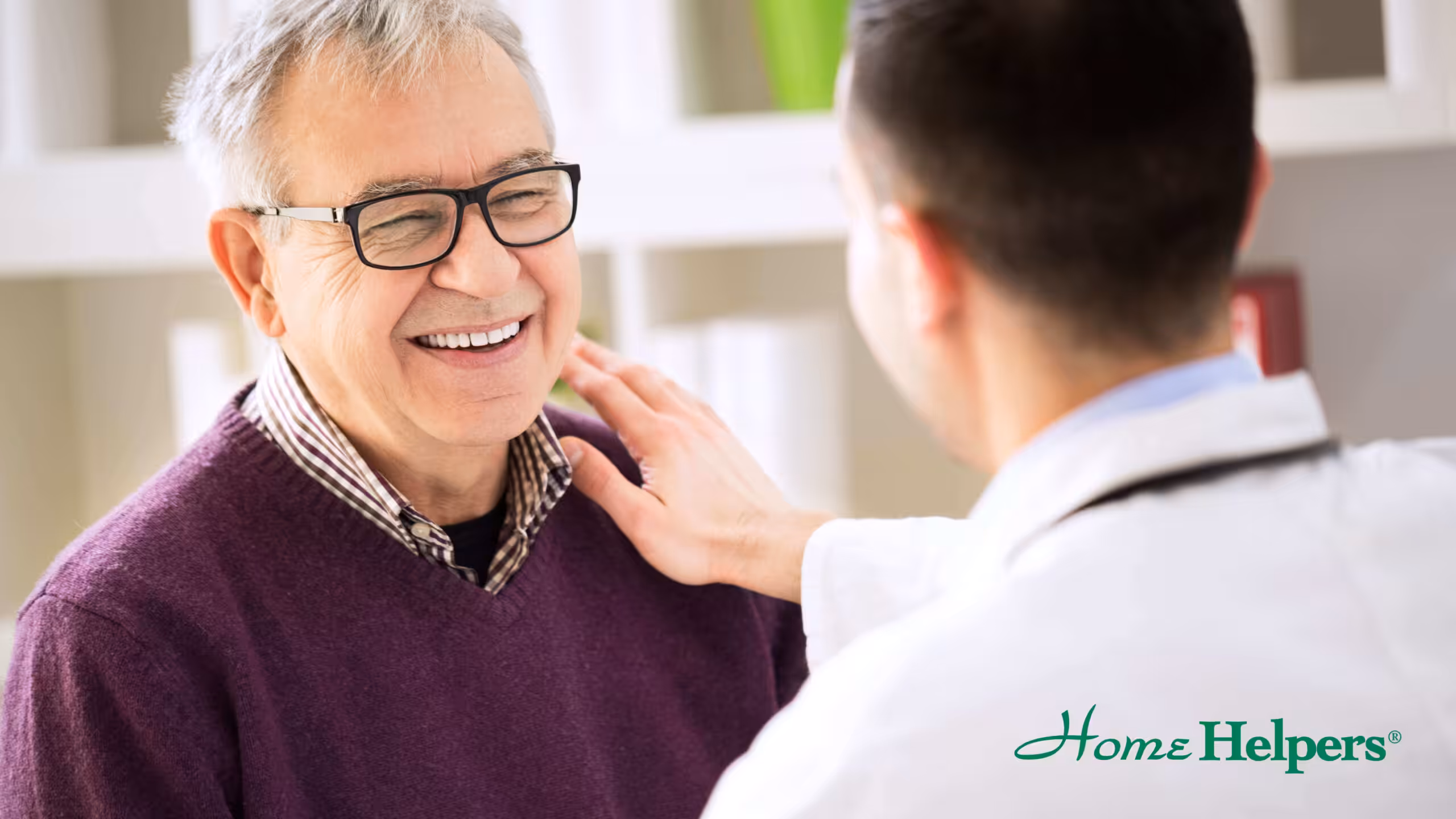 A smiling older man wearing glasses and a purple sweater sits while a healthcare professional in a white coat gently touches his shoulder. The Home Helpers logo appears in the bottom right corner.