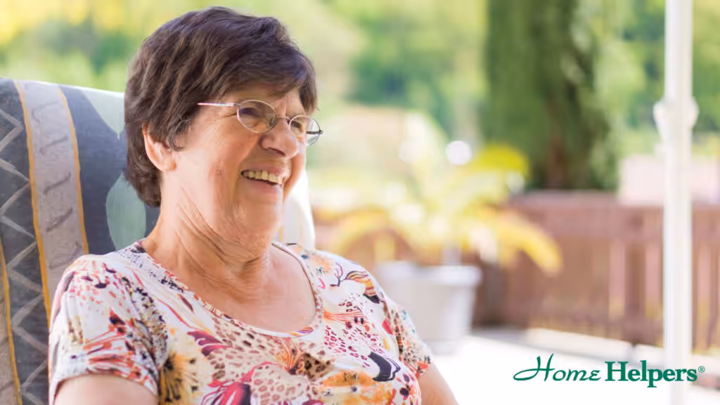 An elderly woman with short brown hair and glasses smiles while sitting outdoors. She wears a floral top, and the background is blurred with greenery. The Home Helpers logo is visible in the lower right corner.
